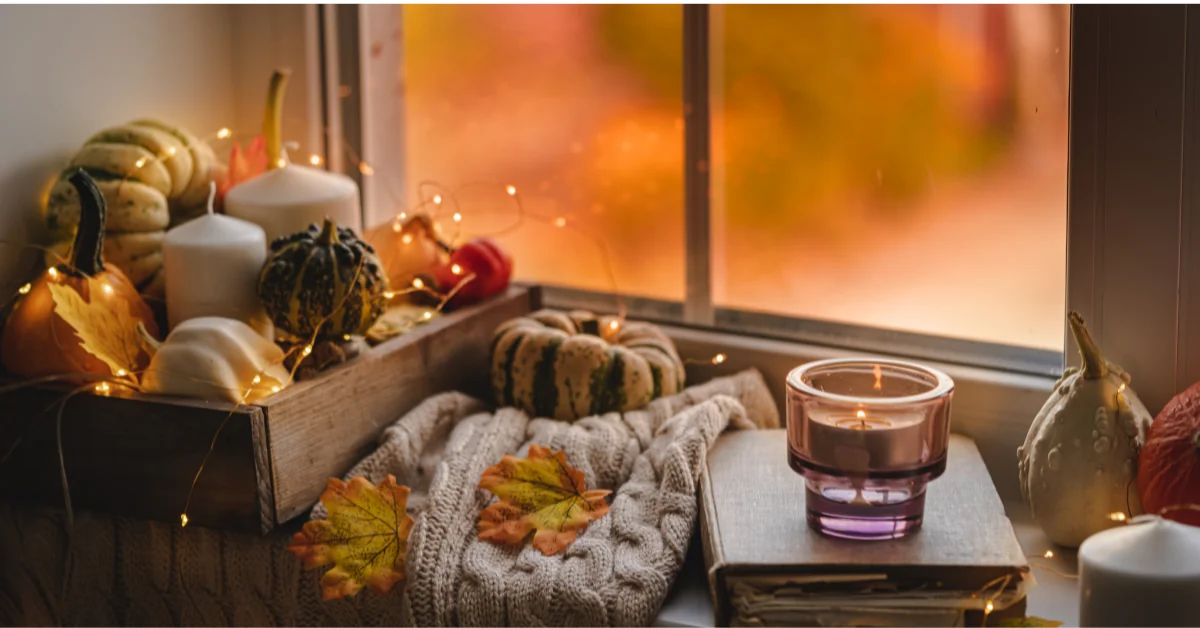 Cozy fall corner with pumpkins in vintage wooden box, candle on designer book, and chunky knit blanket by a window.